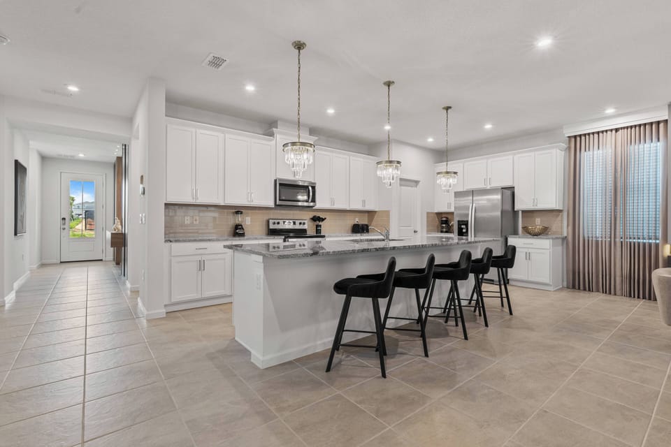 Discover this airy kitchen with a spacious island and sleek black bar stools for casual dining. The warm beige tile pairs beautifully with bright cabinetry, and pendant lights add a soft glow for an inviting atmosphere.