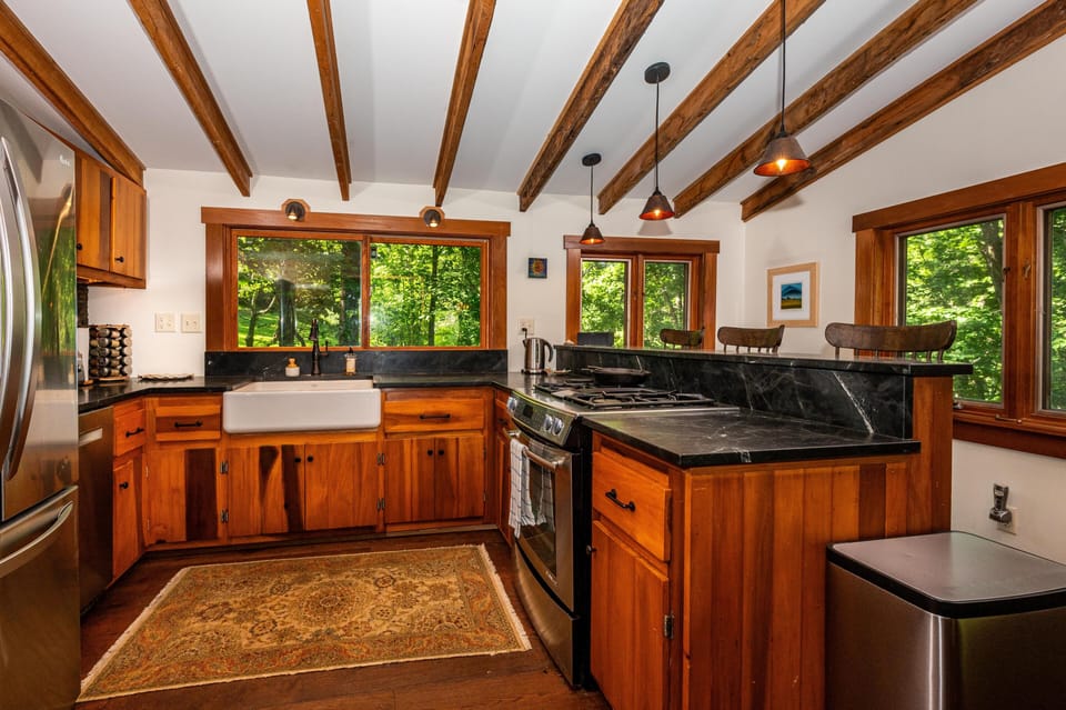 Black granite countertops and a farmhouse sink sit beneath wood-framed windows with forest views in the kitchen.