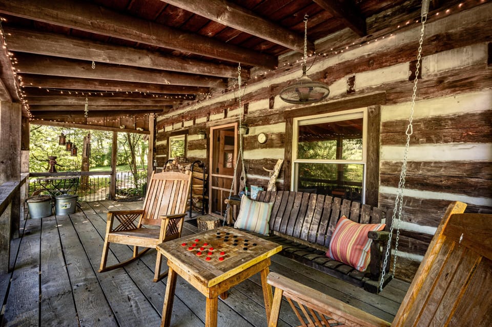Wooden porch with log cabin walls, hanging bench swing, striped pillows, rocking chairs, and checkers table.