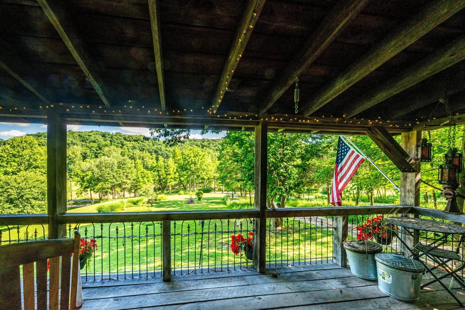 Covered wooden deck with string lights, iron railing, hanging lanterns, American flag, and green hillside views.