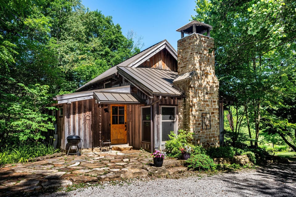 Stone-paved front patio with charcoal grill, weathered wood siding, and a tall stone chimney under a metal roof.