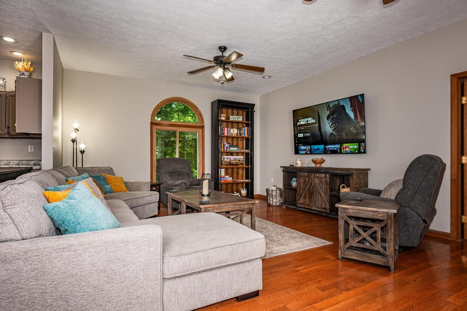 Living room with gray sectional, wooden coffee table, dark recliner chairs, and a flat-screen TV mounted on beige walls.