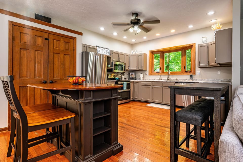 Kitchen features gray cabinetry, stainless steel appliances, and a window with a wood frame overlooking green trees.