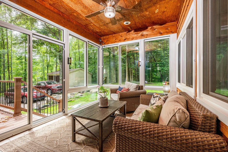 Screened porch with woven rattan sofa and chair, wood coffee table, textured rug, and forest views.
