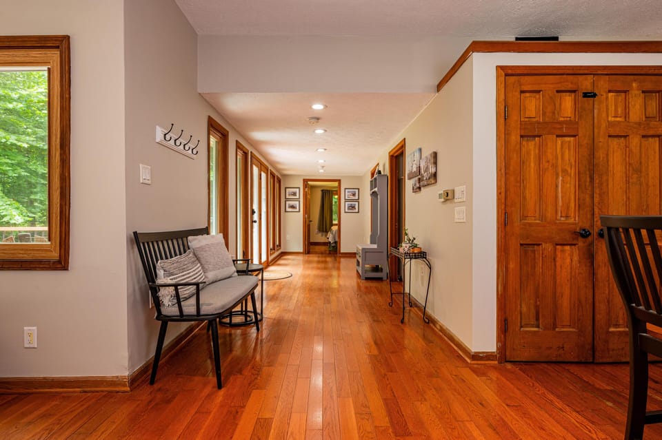 Hallway with polished hardwood floors, black metal bench with gray cushions, and wood-trimmed windows overlooking trees.