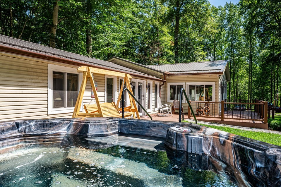 Outdoor hot tub with black and gray marbled finish, wooden swing, and beige vinyl-sided house surrounded by trees.
