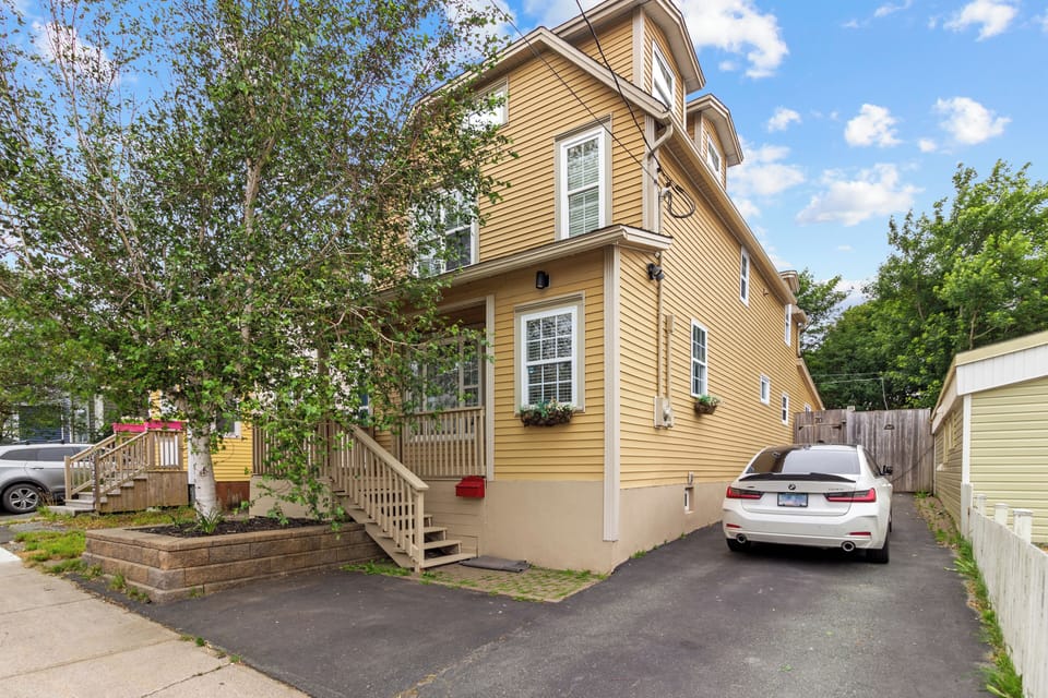 The front exterior of the house features a well-maintained yard, a charming porch, and a driveway with ample parking space. The yellow siding and large windows create a welcoming facade.
