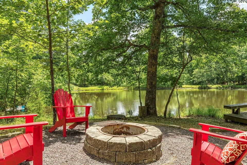 Stone fire pit surrounded by red Adirondack chairs beneath mature trees, just steps from the peaceful lake.