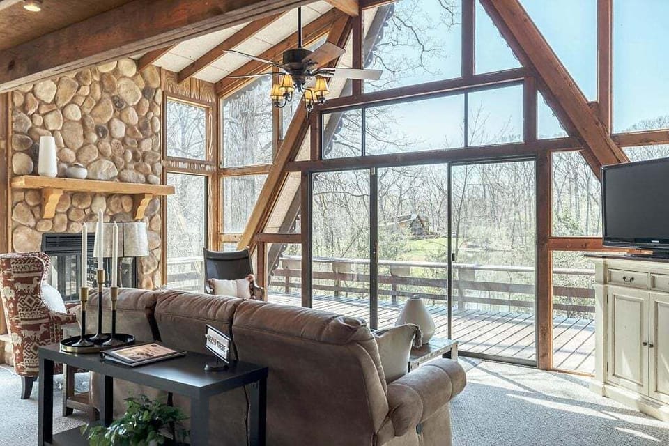 Living room with sofa seating, stone-faced fireplace, TV, vaulted A-frame windows and glass doors overlooking the deck and wooded pond.