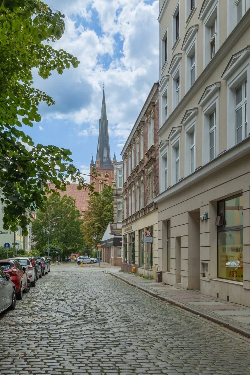 A cobblestone street lined with trees and historic buildings. The entrance to the apartment building is visible on the left.