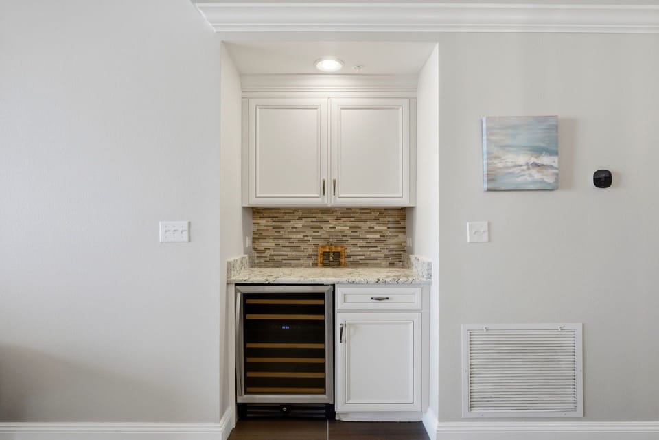 Enjoy coastal elegance in your private wet bar, featuring sleek white cabinetry, granite countertops, a stylish mosaic backsplash, and a chilled wine fridge ready for your evening drinks.