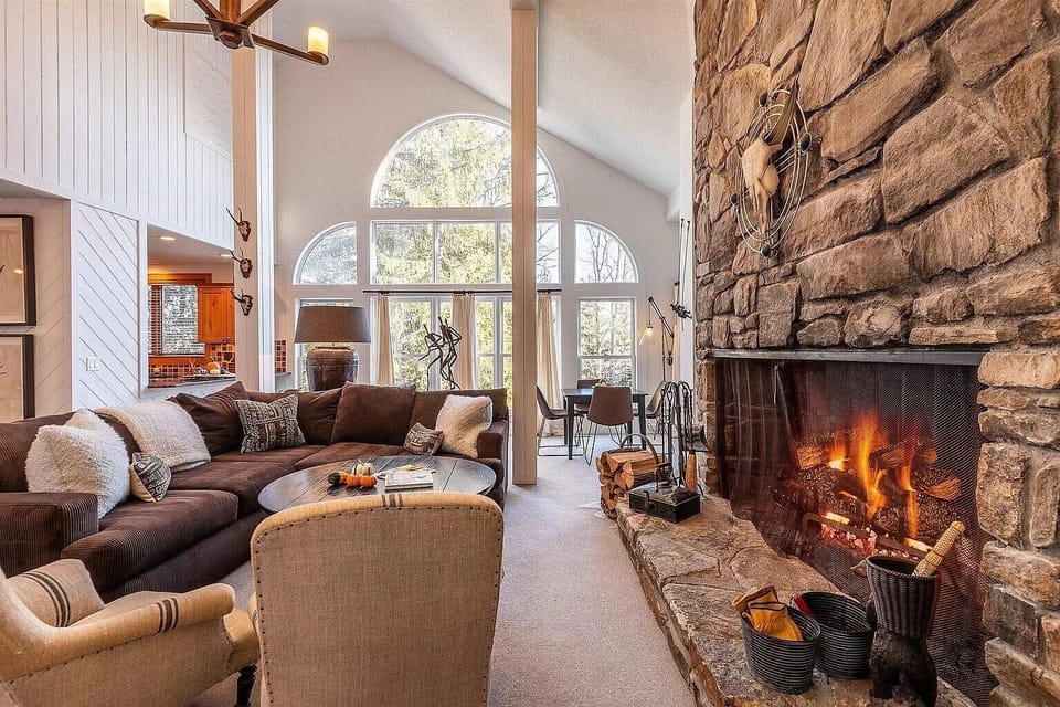 - Cozy mountain living room with stone fireplace and floor-to-ceiling windows   - Relax in rustic charm with forest views and warm lighting   - Perfect ski cabin vibe for your mountain getaway