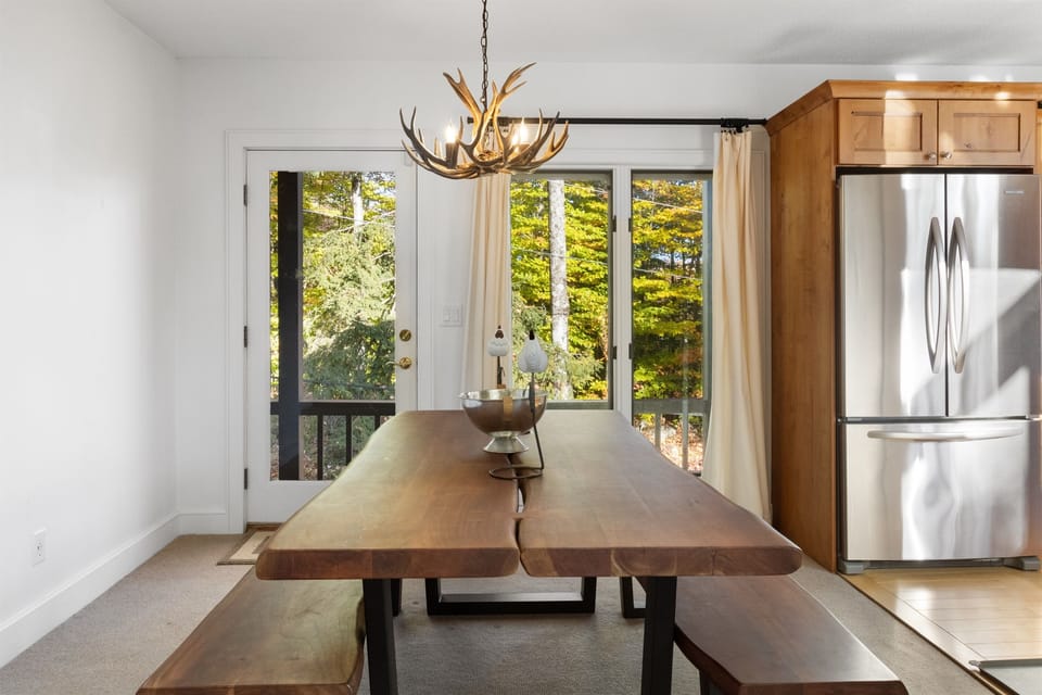 - Rustic dining area with live edge wood table and antler chandelier   - Natural light and forest views through sliding glass doors   - Perfect space for family dinners after mountain fun