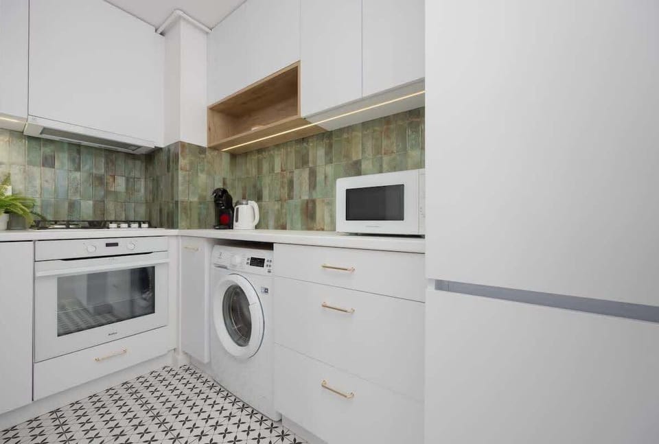 A compact kitchen with white cabinets, a washing machine, microwave, and tiled backsplash. The layout is practical and modern for everyday cooking.