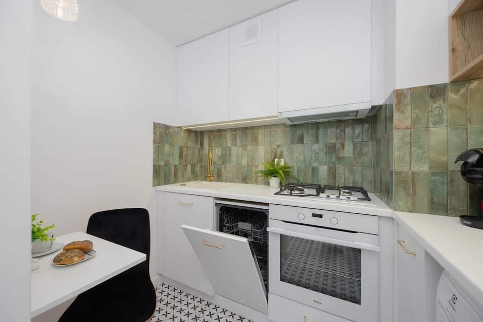 A white kitchen with a tiled floor, green backsplash, and compact layout. The space includes a stove, oven, and small dining area.