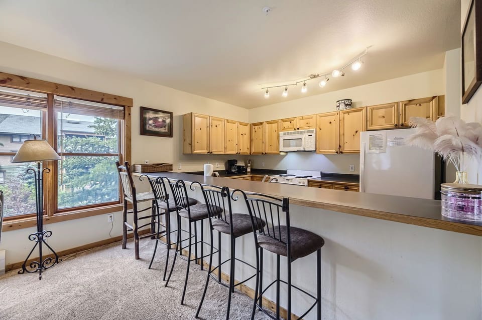 A kitchen with wooden cabinets, a bar counter with four chairs, a microwave, and a refrigerator. Track lighting illuminates the space, and there is a window with blinds on the left side.