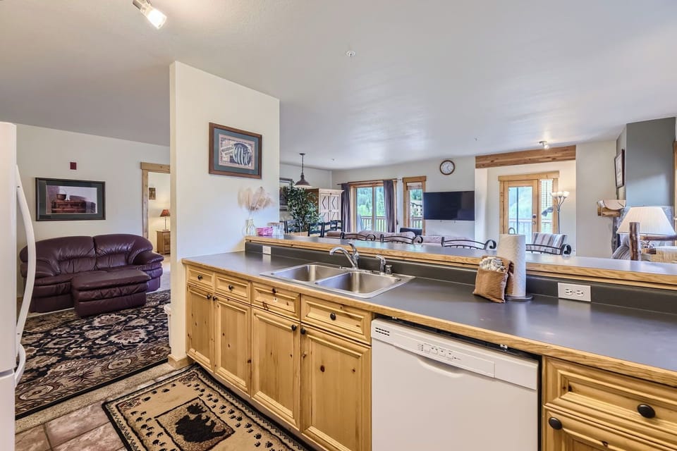 A kitchen with a double sink, dishwasher, and wooden cabinets facing an open living room with seating, a TV, and large windows.