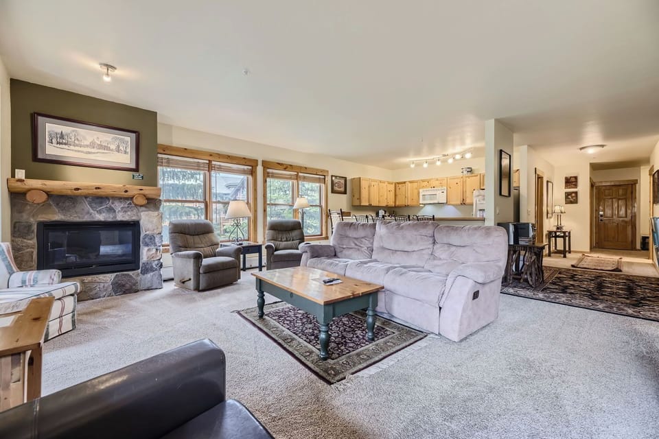 A cozy living room with a stone fireplace, large windows, a beige sectional sofa, a wooden coffee table, and an open kitchen in the background with light-colored cabinetry.