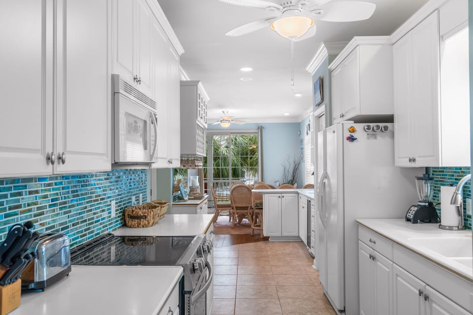 Your bright galley kitchen features gleaming white cabinets, stunning turquoise backsplash, and flows seamlessly to your dining space with tropical views.