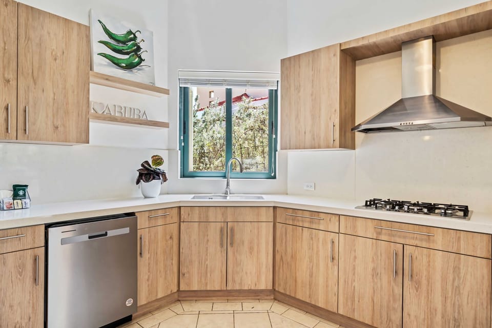 Sleek wooden cabinets and a stainless steel range hood highlight this modern kitchen - A window allows natural light to brighten the space, featuring green-themed artwork - The kitchen includes a built-in dishwasher and a compact sink for ease