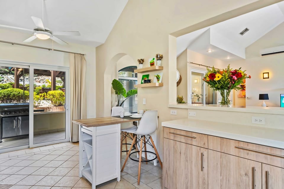The kitchen features a cozy breakfast bar with stylish white stools for casual dining - Glass doors open to an outdoor area with a barbecue grill, perfect for entertaining - Bright flowers and open shelves add a touch of charm to the place
