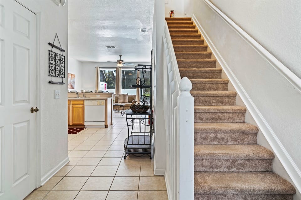 Open living area with a view of the kitchen and cozy seating - Carpeted staircase leading to the upper level, adding a touch of warmth and comfort - Functional kitchen space with essential appliances and a clear sightline to the living room