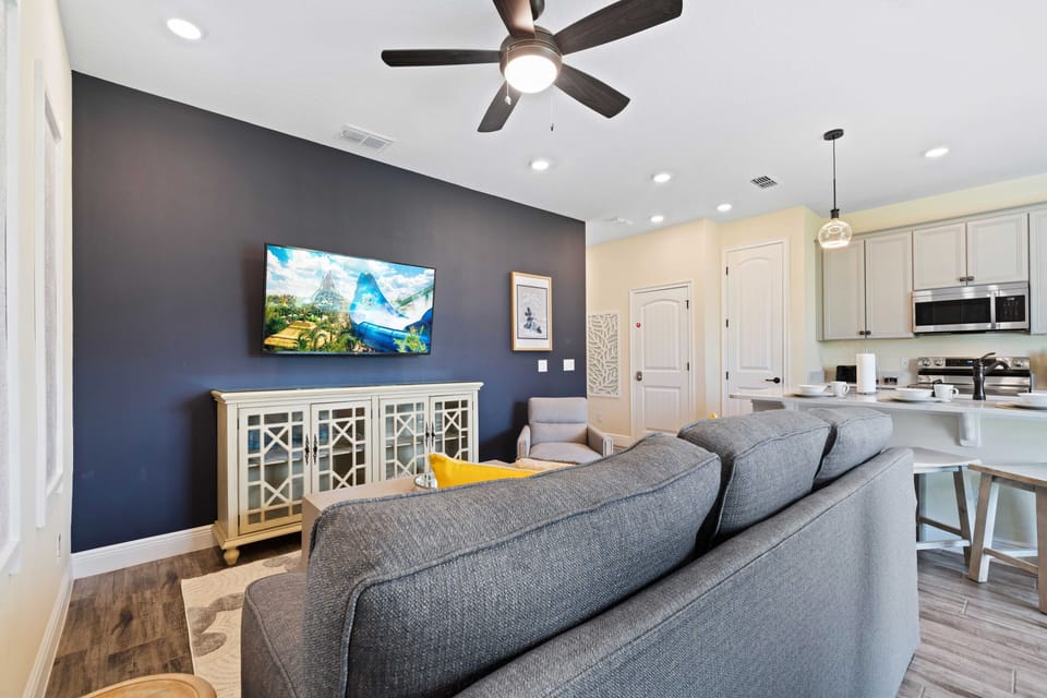 Bright and welcoming living room with a plush gray sofa and a stylish armchair - Navy accent wall features a mounted flat-screen TV above a vintage-style cabinet - Open floor plan flows into a contemporary kitchen and a cozy breakfast bar
