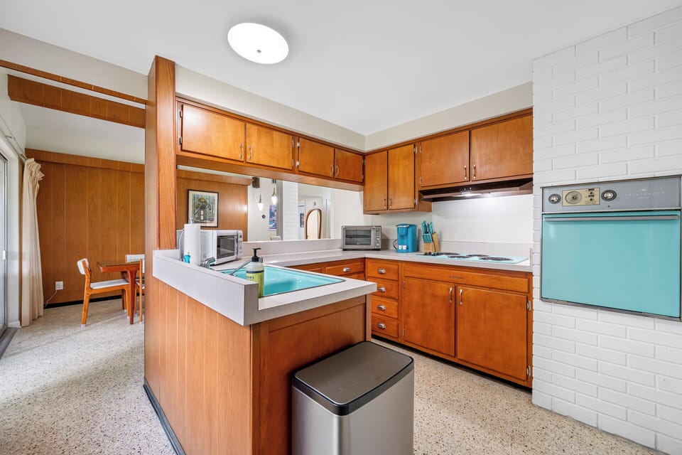 Retro mid-century kitchen with warm wood cabinetry, turquoise appliances, terrazzo floors, and a bright open layout connecting to the dining area.