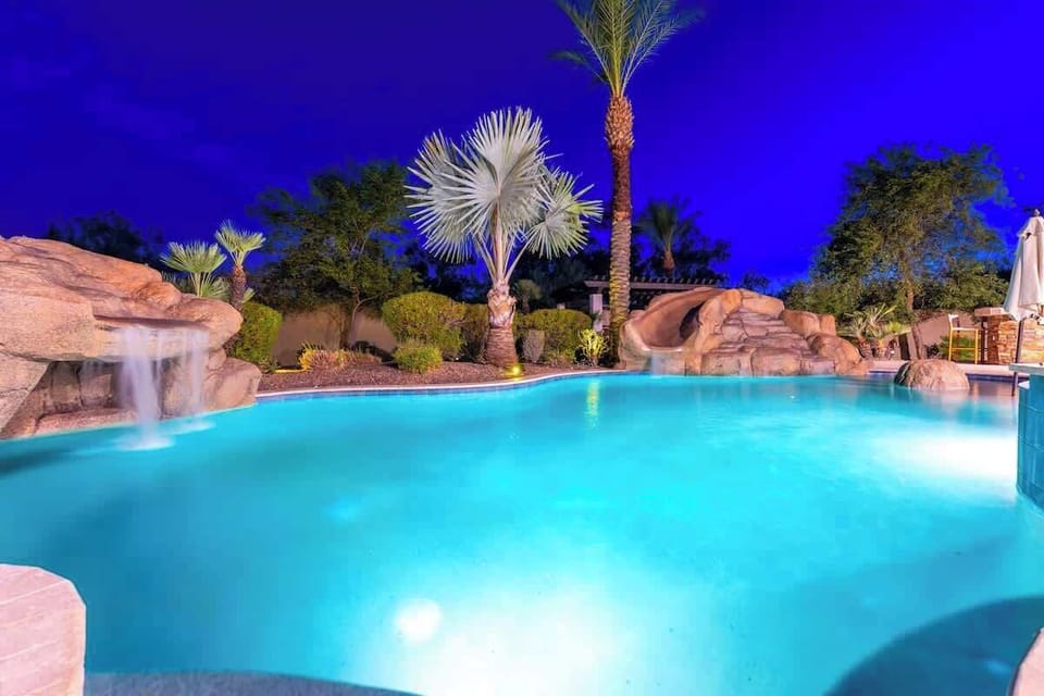 Picture yourself gliding into crystal-clear waters under a vibrant desert sky. This stunning pool features natural rock waterfalls, tropical palms, and a serene evening glow—your perfect escape awaits.
