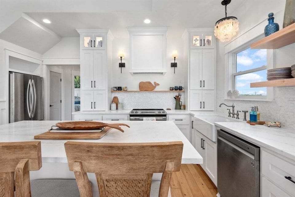 White coastal kitchen with stainless steel refrigerator, range hood, island prep space, and bright natural light.