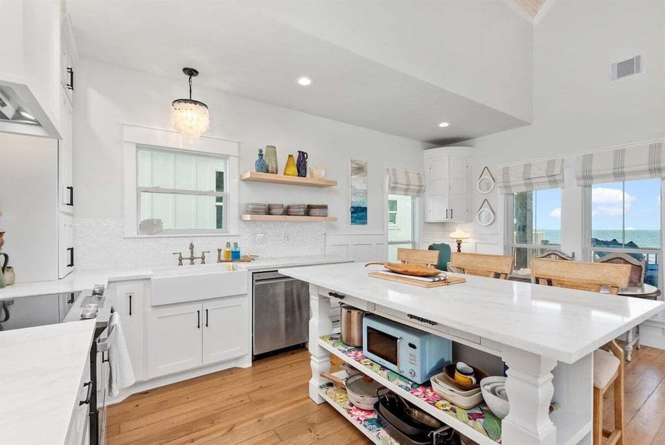 Bright kitchen view with white cabinetry, island storage, dishwasher, open shelving, and coastal wood floors.