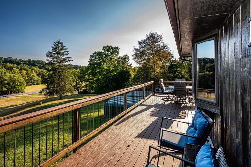 Long wood deck with black metal railings features cushioned chairs and a round dining table overlooking grassy hills and trees.
