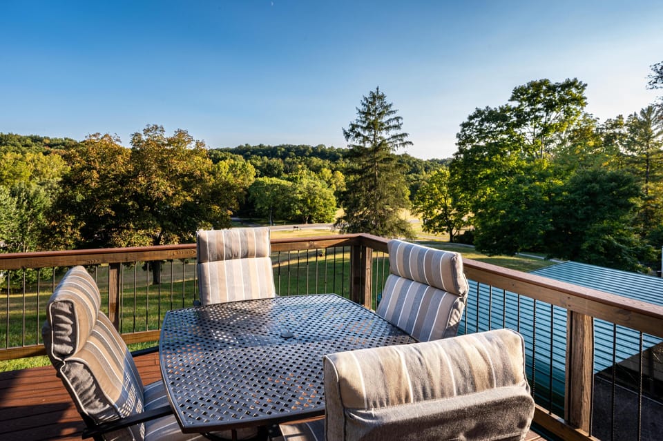 Outdoor deck with metal dining table and striped cushioned chairs overlooking green trees and rolling hills.