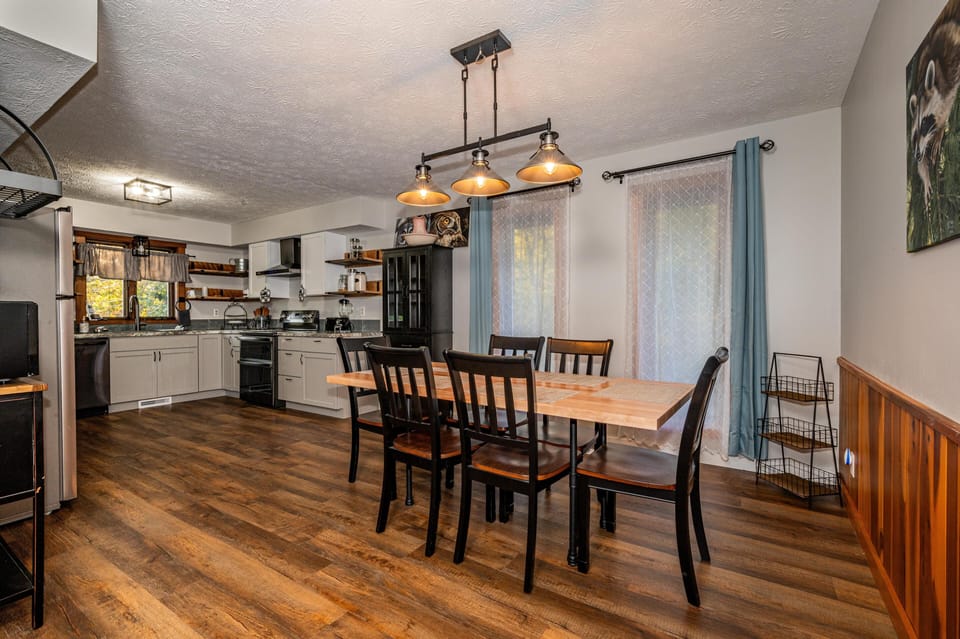 Dining area with wood table and black chairs beneath industrial-style pendant lights by sheer curtained windows.