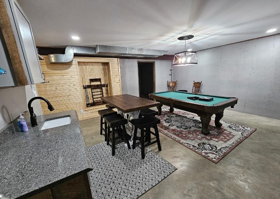 Basement game room with green felt pool table on patterned rug, wood-top table, and granite countertop wet bar.