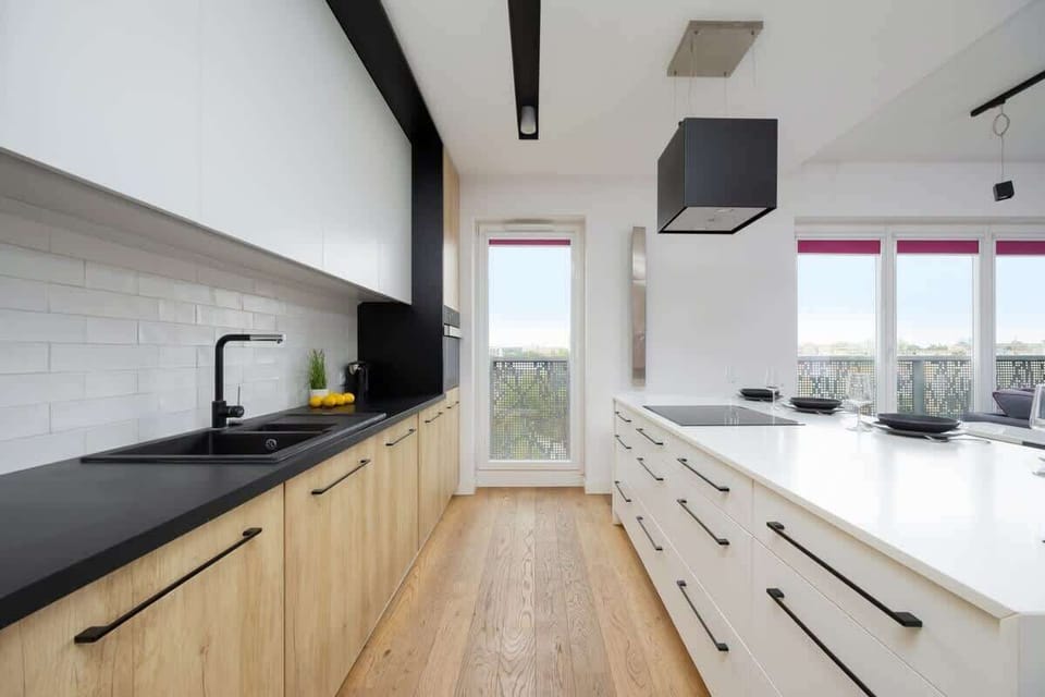 A minimalist kitchen with light wood cabinetry and clean white countertops. Large windows bring in plenty of daylight.