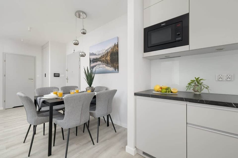A modern kitchenette with a dining table set for four. Clean white cabinets and large windows fill the space with light.