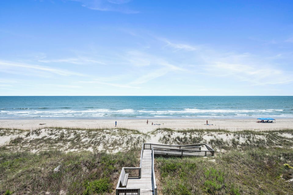 A private boardwalk leads over sandy dunes directly to the pristine oceanfront, with calm waves and open sky stretching beyond.