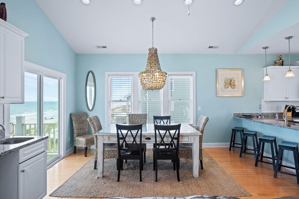 Gather at the dining table with ocean breezes and sunlight framing every shared meal, while the elegant chandelier and coastal accents create unforgettable moments in your beach retreat.