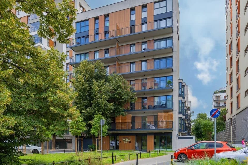 A modern apartment building with a tree-lined sidewalk and parked cars. The facade features large windows and clean architectural lines.
