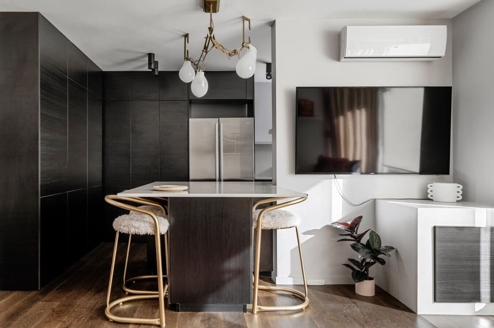This stylish kitchen island doubles as a breakfast bar with plush gold-accented stools, set beneath a sculptural light fixture and adjacent to a large smart TV for relaxed dining and entertainment