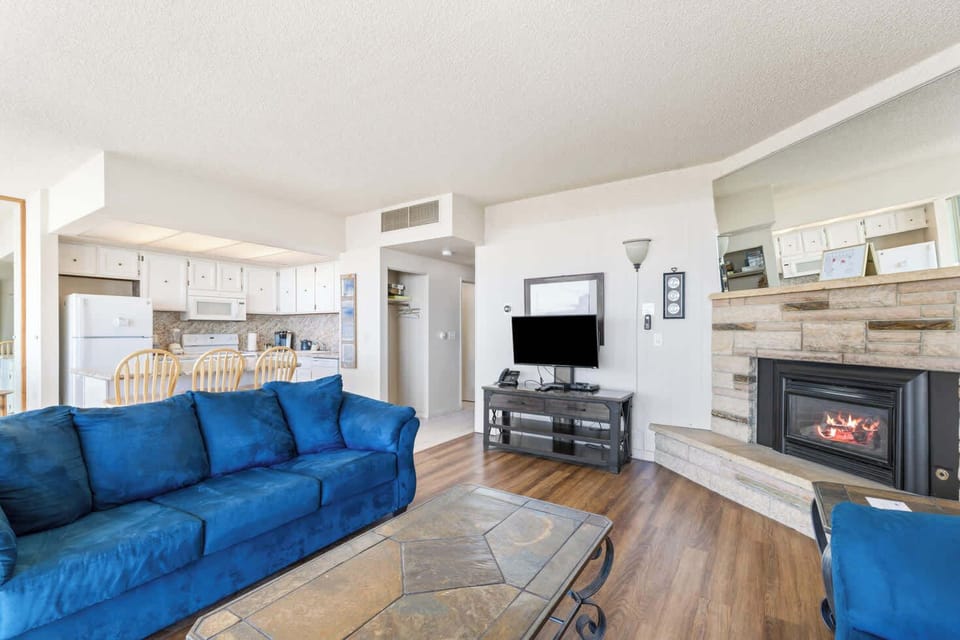 This inviting open-concept living space features a cozy fireplace, comfortable sofas, a flat-screen TV, and a glimpse of the well-equipped kitchen.