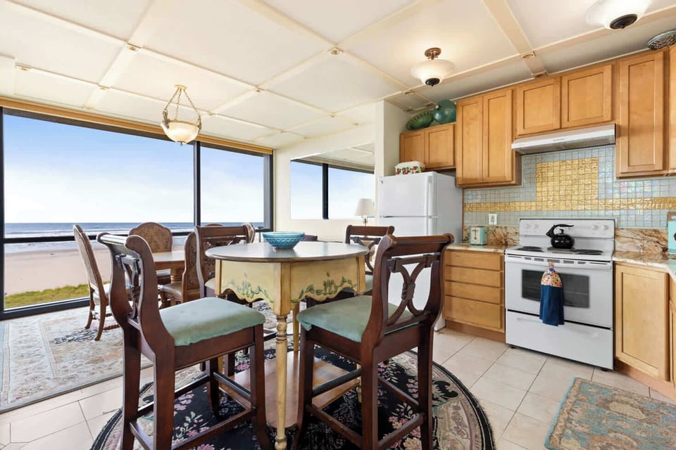 Oceanfront dining with a view! This open-concept space features a well-equipped kitchen, a cozy dining table, and expansive windows showcasing stunning beach and sea scenery.