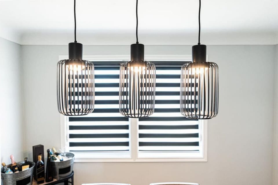 `Illuminate your dining experience in our stylish dining area 1. Three modern pendant lights with unique caged designs provide ample, warm lighting. Behind them, a large window fitted with sleek horizontal blinds offers privacy and natural light. Enjoy th