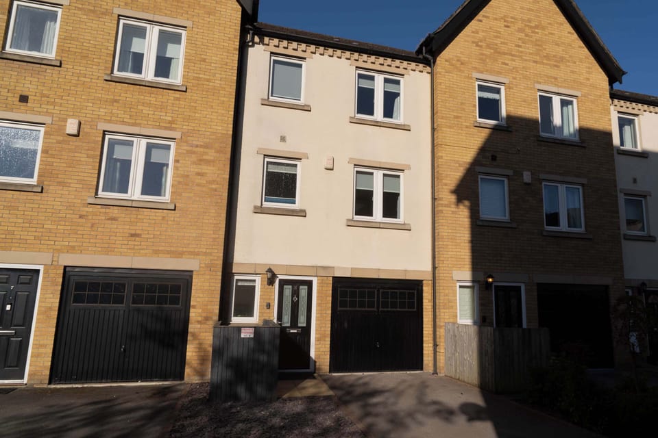 An exterior view of a modern three-storey townhouse with a neutral facade and integrated garage. The property is neatly positioned between two brick-fronted homes, with crisp sunlight casting clean shadows on the building.
