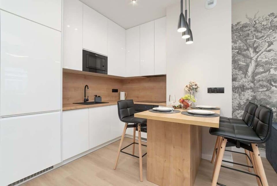 A modern kitchen with white cabinets, wood accents, and a dining area. The space is open and bright with a clean, minimalist design.