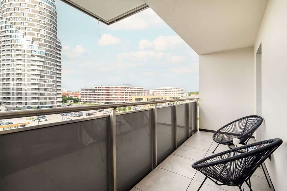 A balcony view with black chairs and a table, overlooking modern city buildings. The railings are glass for an unobstructed view.