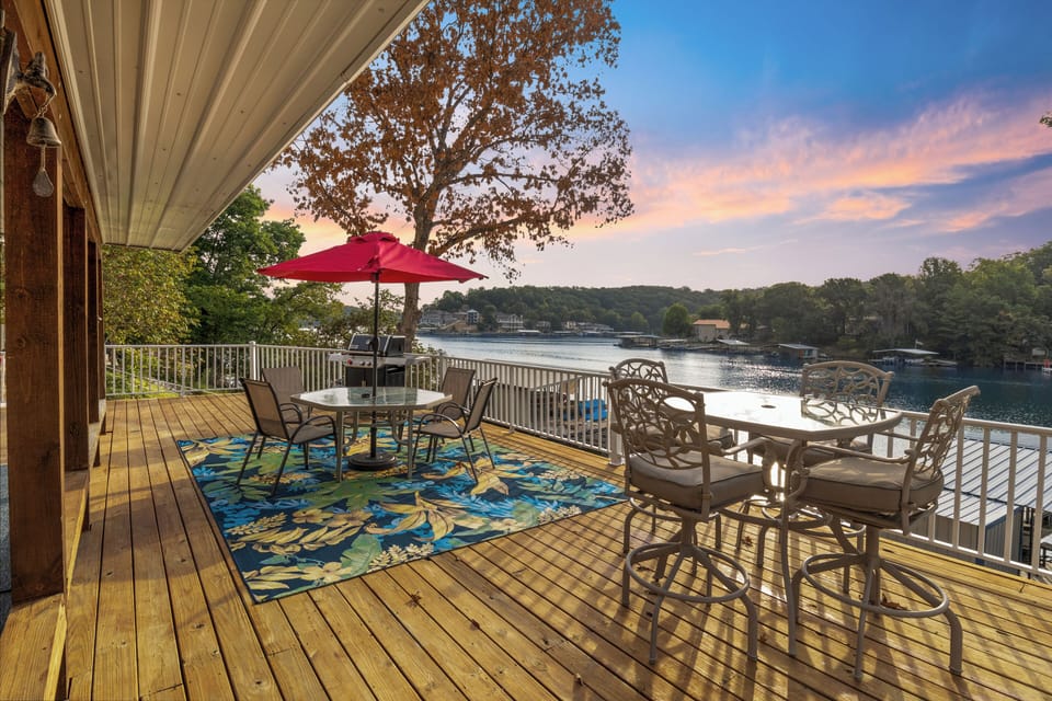 Lakeside deck with stunning sunset views, cozy seating, vibrant decor, and a BBQ grill—perfect for relaxing or dining.