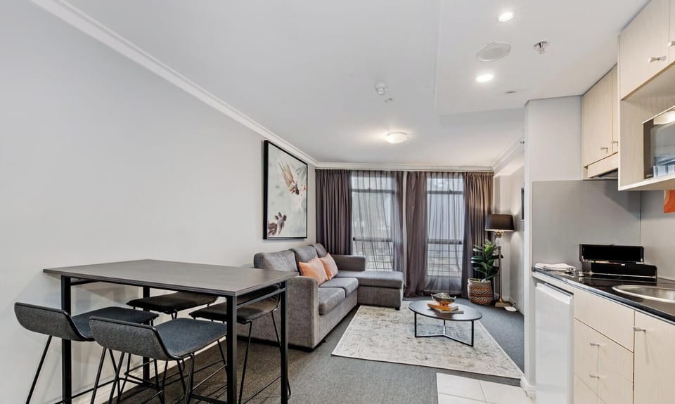 A perspective showing a bar-height dining table with stools, leading past the open kitchenette toward the sofa area in the back.