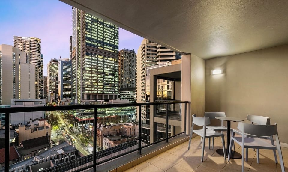 An outdoor balcony area at dusk, furnished with a dining table and chairs, overlooking illuminated city buildings.